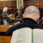 A chapel setting with one student leading a group of students and staff in morning prayer. 'Common Worship: Morning Prayer on a Saturday' is open in the foreground.