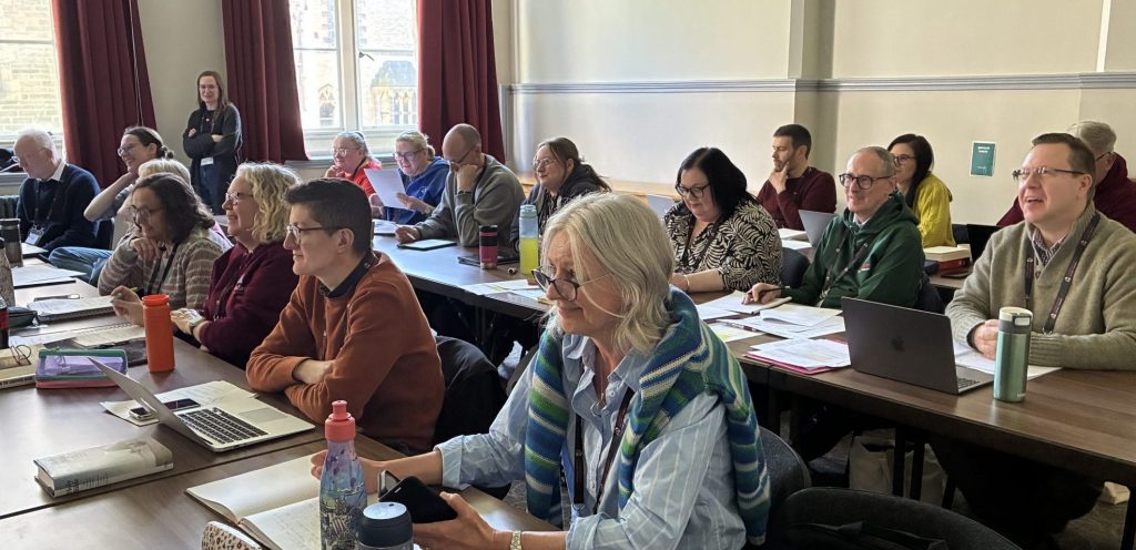 Students sit in three rows in a classroom during a teaching session. Laptops, papers, coffee cups and water bottles cover the tables.